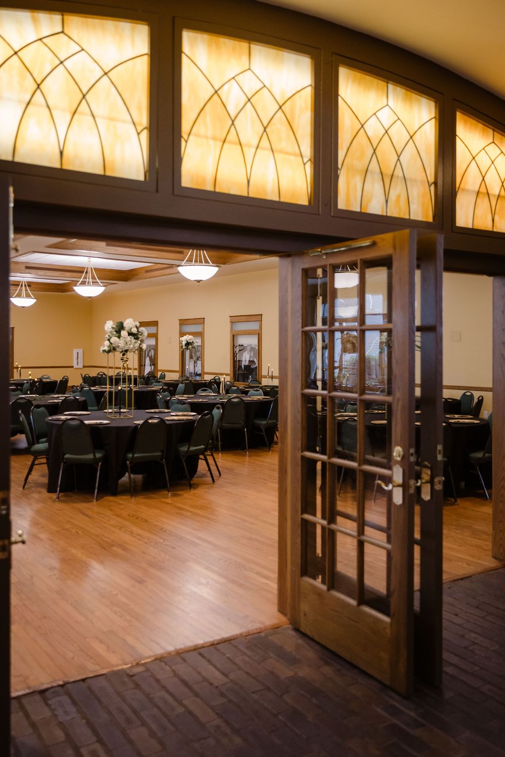 Elegant banquet hall with wooden French doors, hardwood floors and illuminated Art Deco transom windows above.