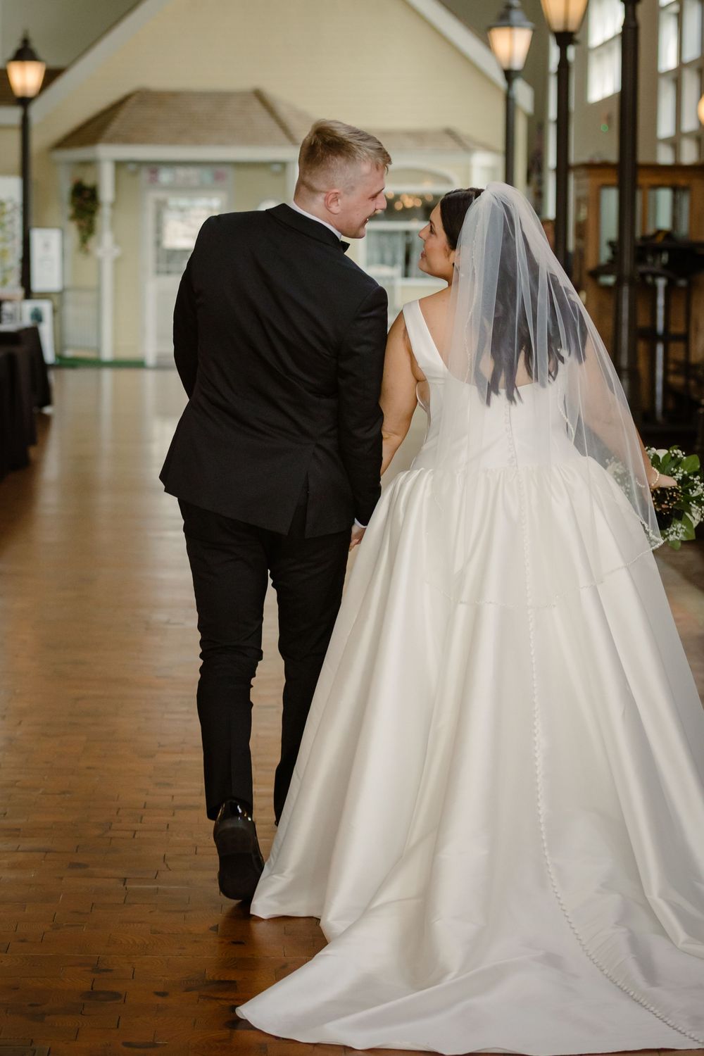 A bride and groom walk together down a hallway with wooden floors and lanterns on their wedding day.