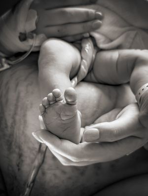 Förlossningsfotografering på Karolinska Solna. Näbild på en fot som ligger i mammans hand med navelsträngen ringlandes bakom
