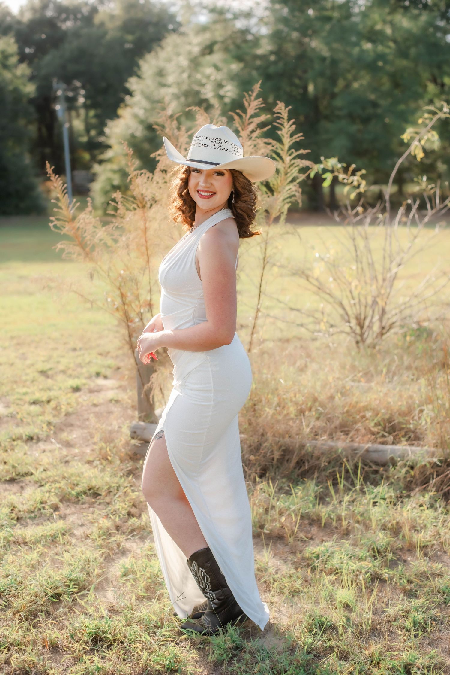 Person in a white halter dress and straw hat poses outdoors in a sunlit natural setting.