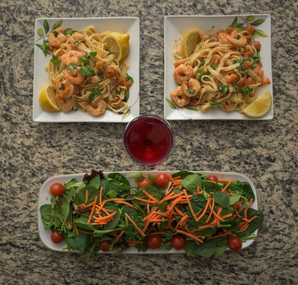 Elegant plating of shrimp scampi dishes with fresh salads and garnishes on a granite countertop with a red beverage.