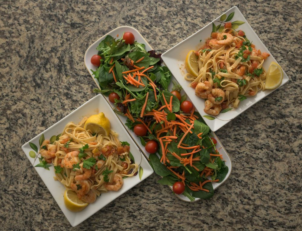 Overhead view of pasta dishes with shrimp and a fresh green salad with carrots served on a granite countertop.