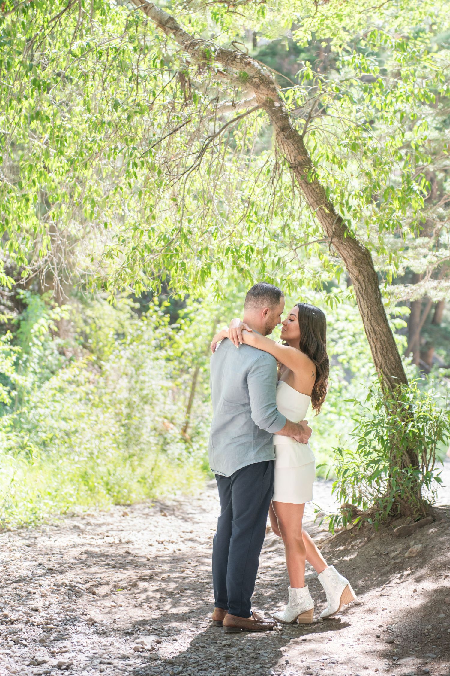 An eloping ouple embracing and almost kissing under a lush green tree on a sunny day outdoors.