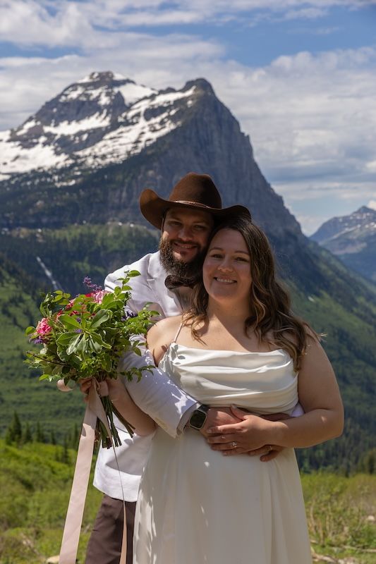 Couple poses in wedding attire with mountain backdrop, groom in cowboy hat hugging bride holding pink bouquet.
