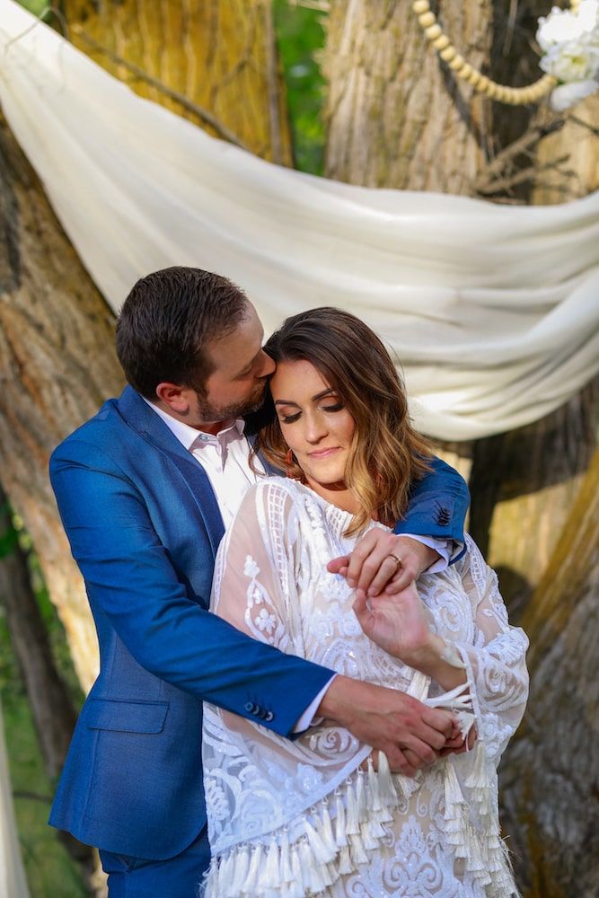 A romantic wedding couple embraces under flowing white fabric draped between trees in an outdoor setting.