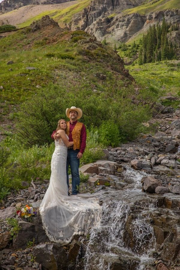 A couple poses together near a cascading mountain waterfall surrounded by lush greenery and rocky terrain.
