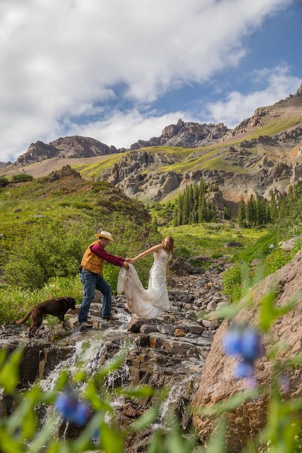 Hiker in orange jacket stands on rocky mountain terrain with purple wildflowers and rugged peaks in the background.