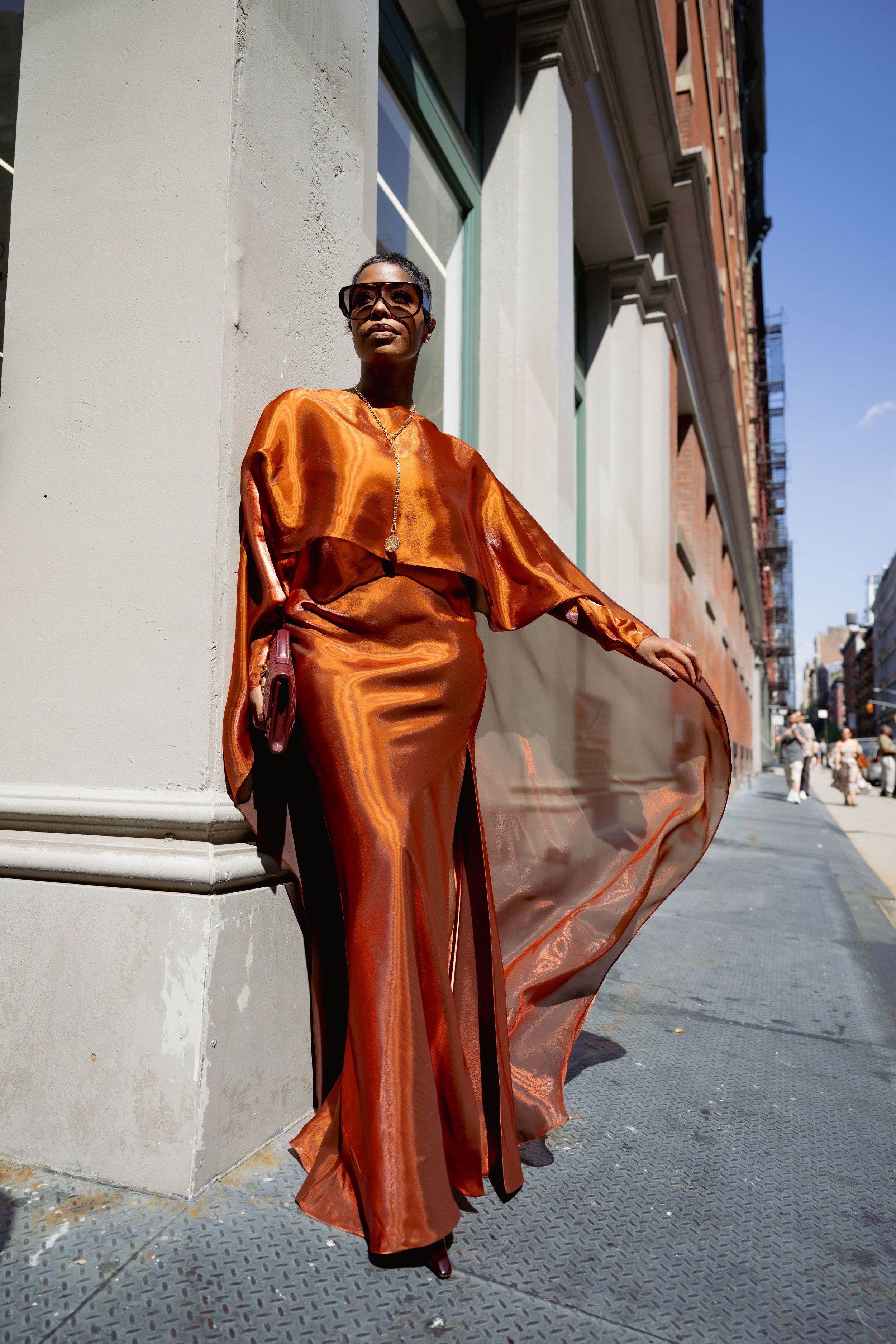 A person wearing a flowing copper-colored satin dress poses dramatically against a white building wall on a city street.