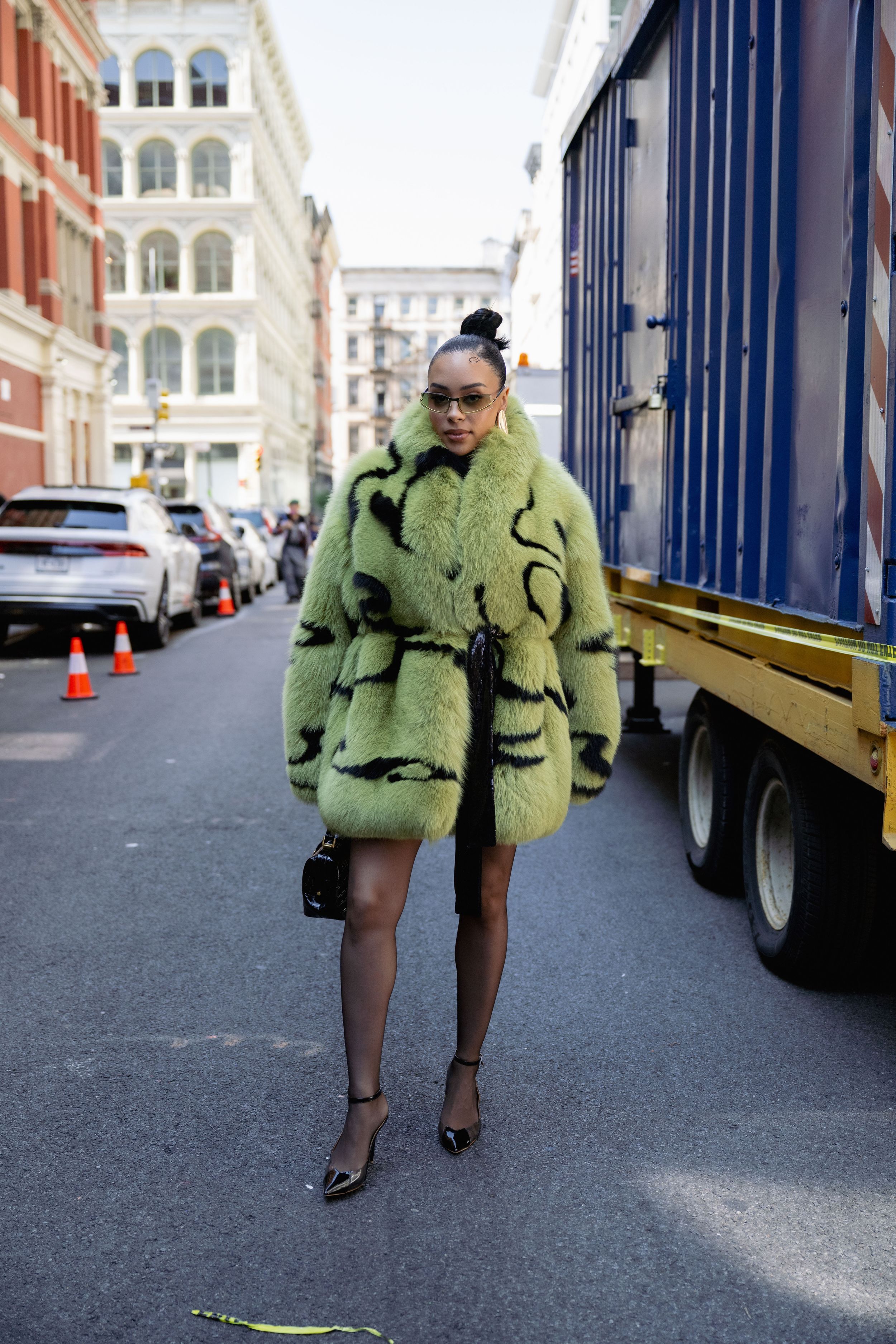 A person wearing a green furry coat and black heels walks past a delivery truck on a city street.