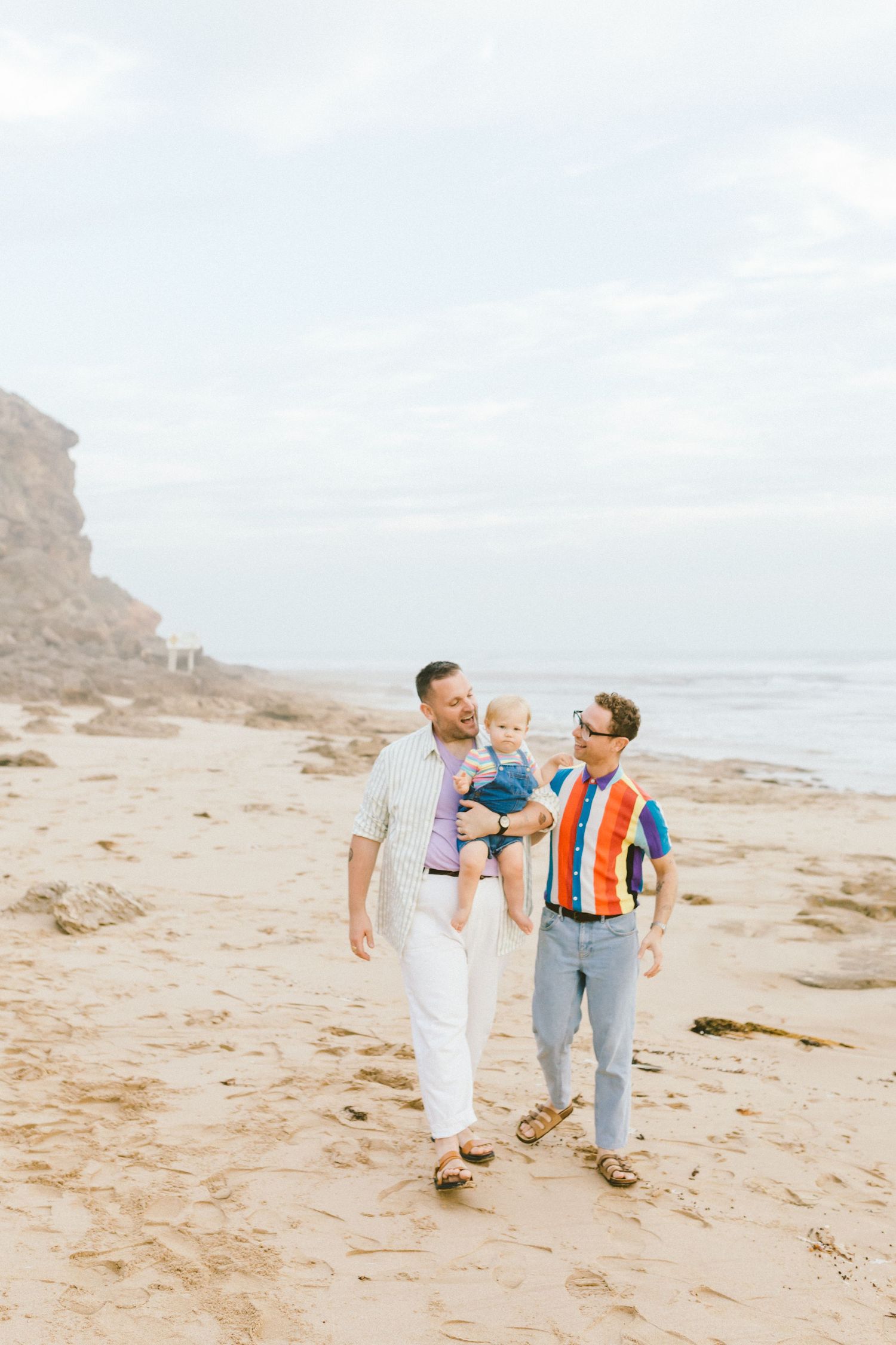 Three people walking and laughing together on a sandy beach with cliffs and ocean waves in the background.