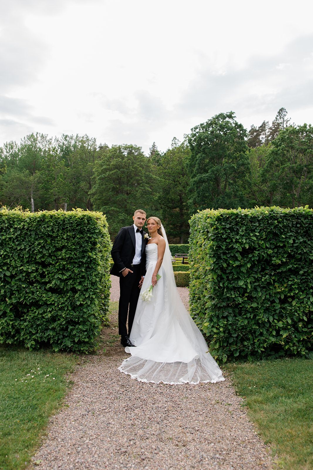 Couple in wedding attire walking along a gravel path bordered by tall manicured hedges in a garden setting.