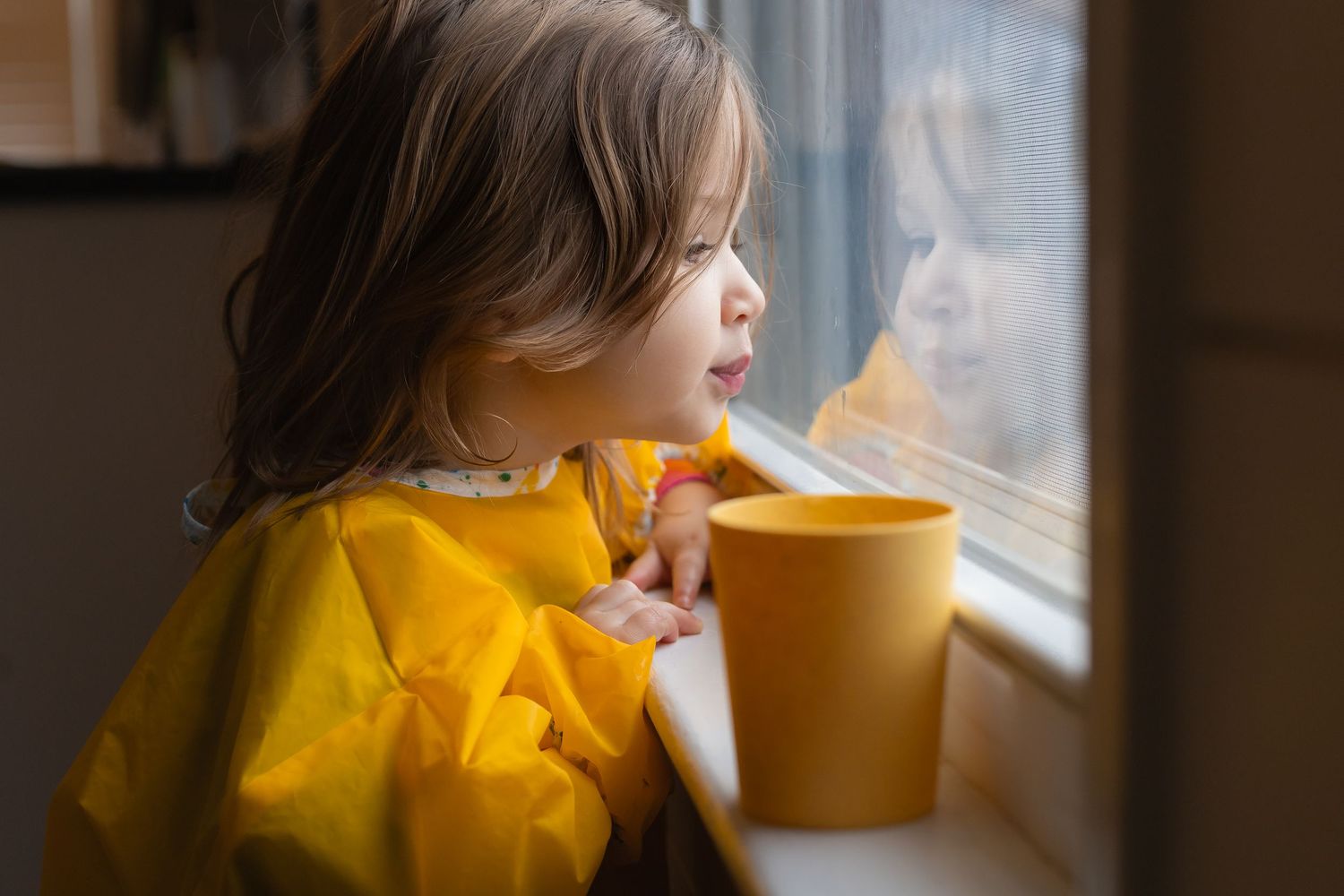 Sioux City, Iowa photographer. Young toddler girl in yellow bib looking out window while holding a yellow cup on a rainy day.
