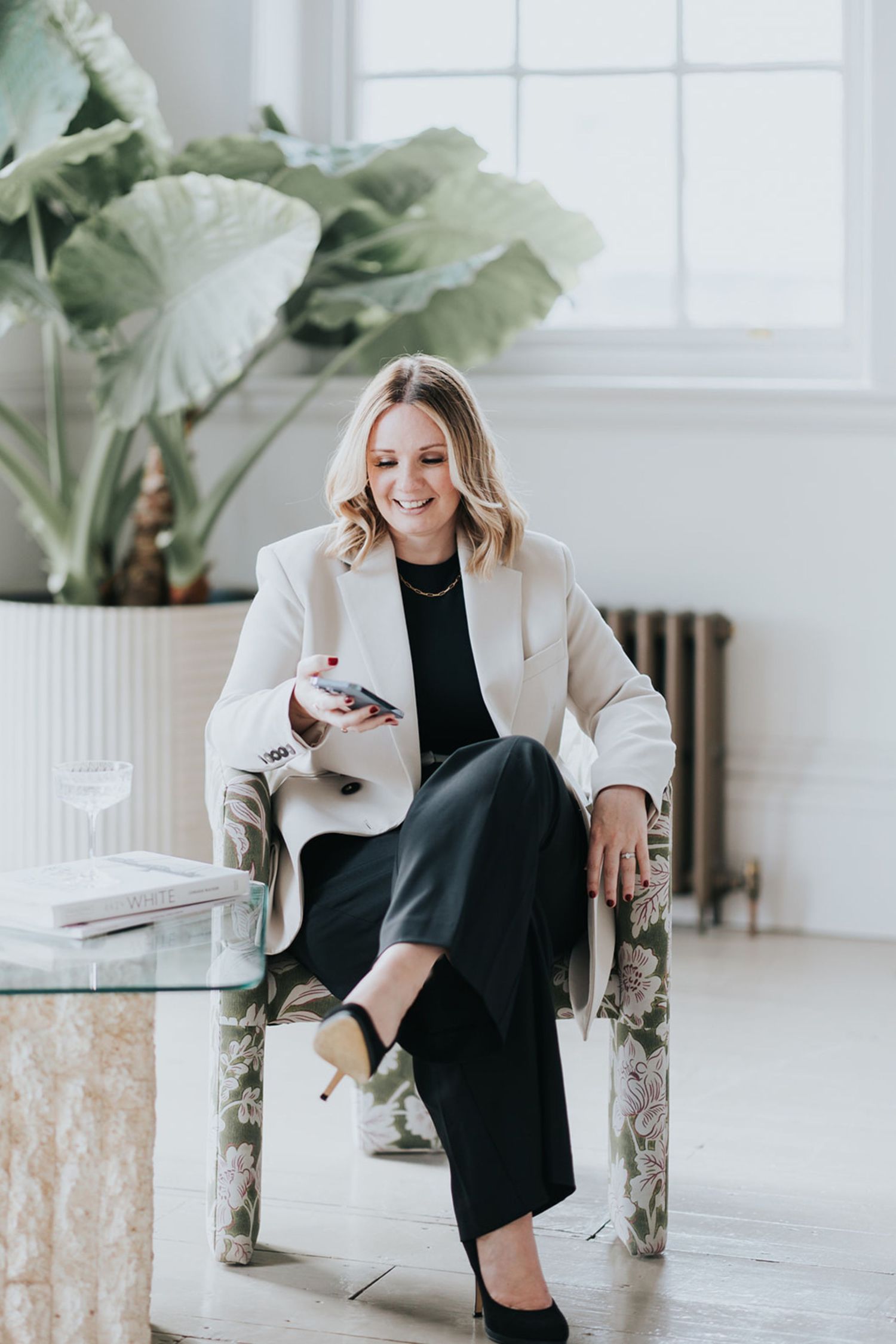 Professional lifestyle photos of woman in black dress and beige blazer posing on modern chair in bright white room.