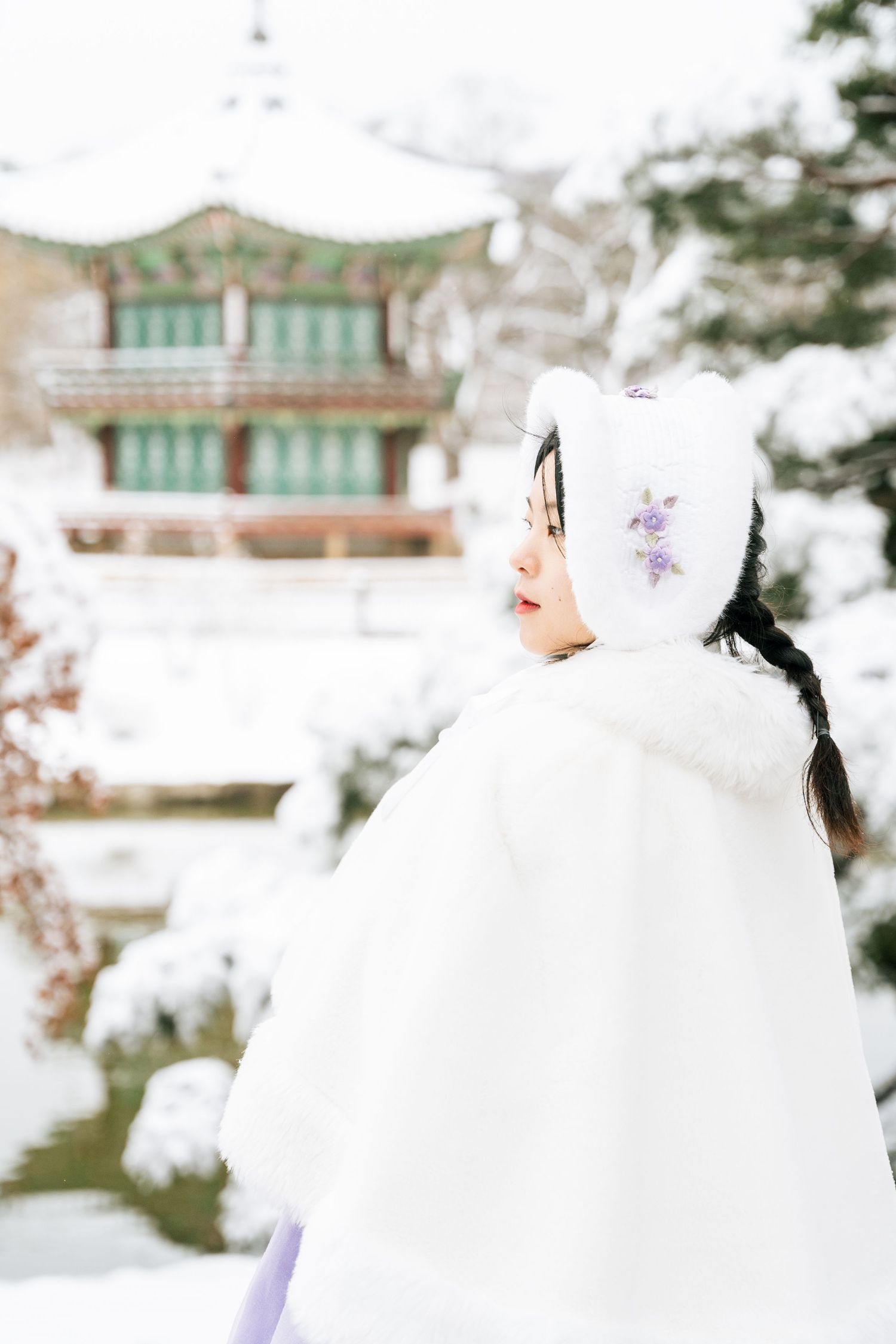 Two figures in traditional white Korean hanbok dresses stand in a snowy winter garden with traditional buildings visible.