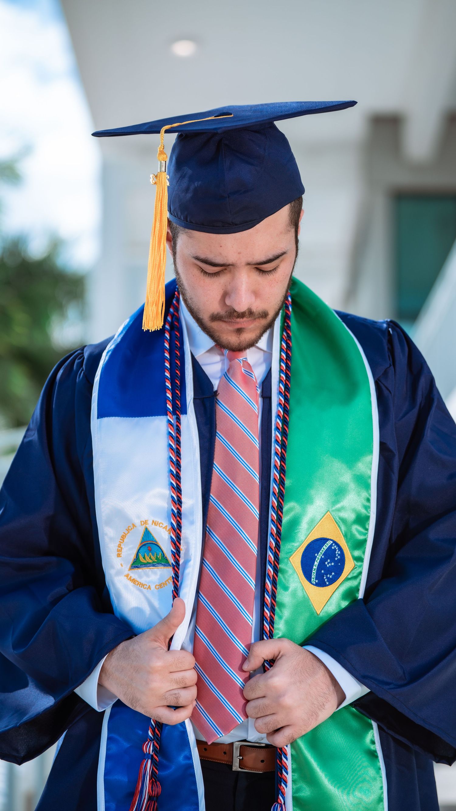 Graduate wearing navy cap and gown with Nicaragua and Brazil flag sashes adjusts tie during commencement ceremony.