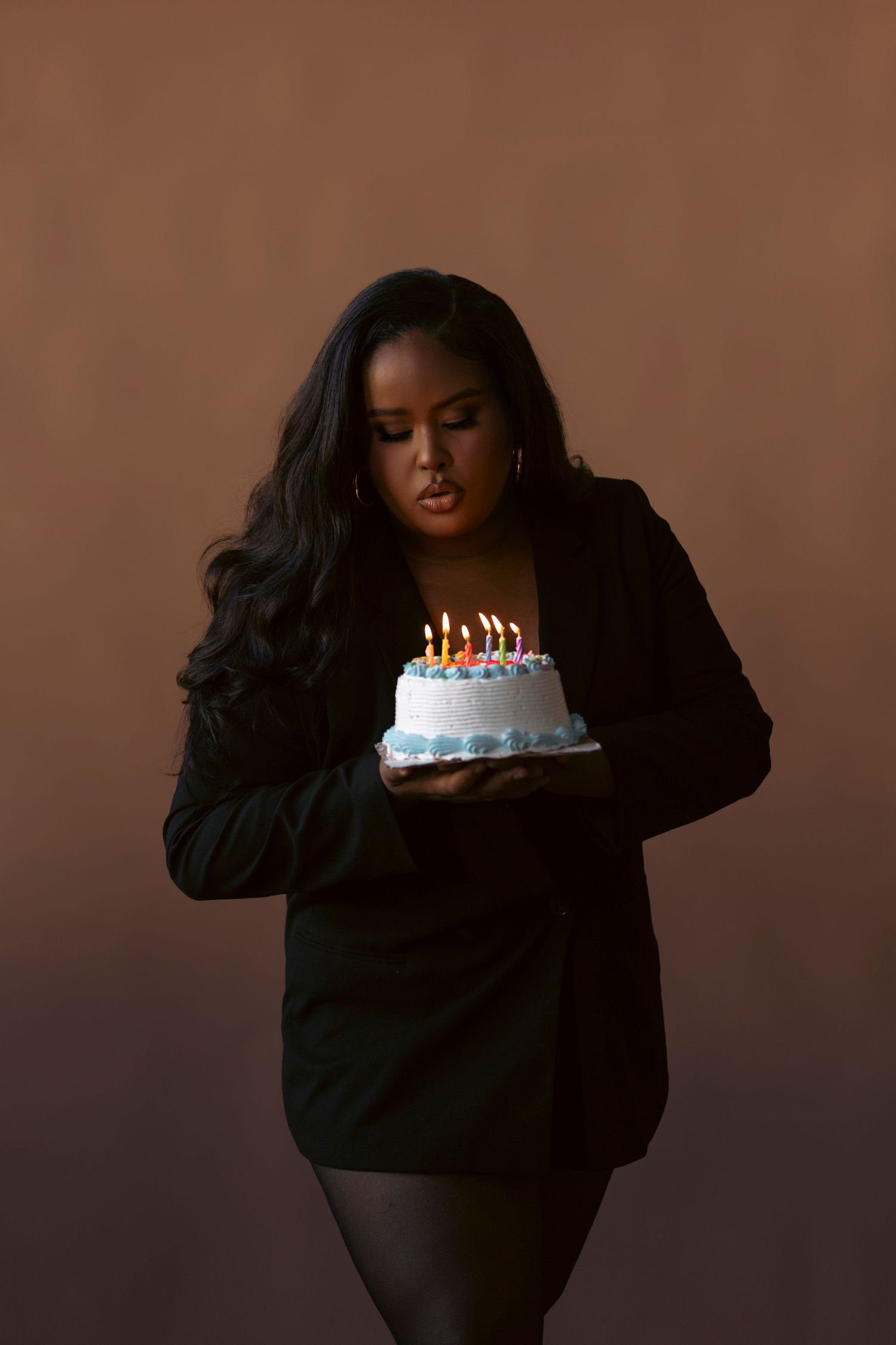 A person in black clothing holds a birthday cake with lit candles against a dark brown background.