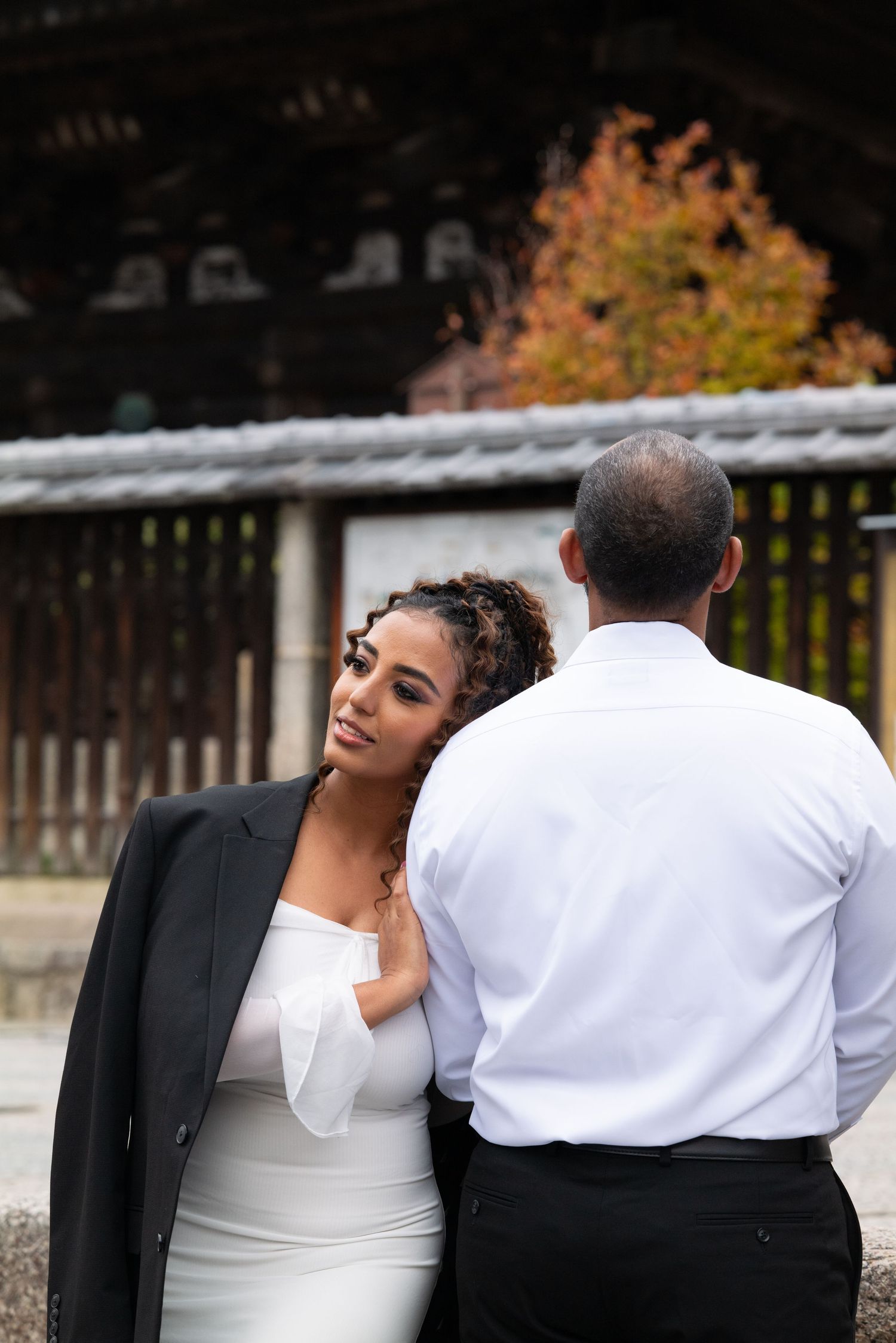 Bride in white off-shoulder dress and black blazer leans on groom at a Japanese temple.