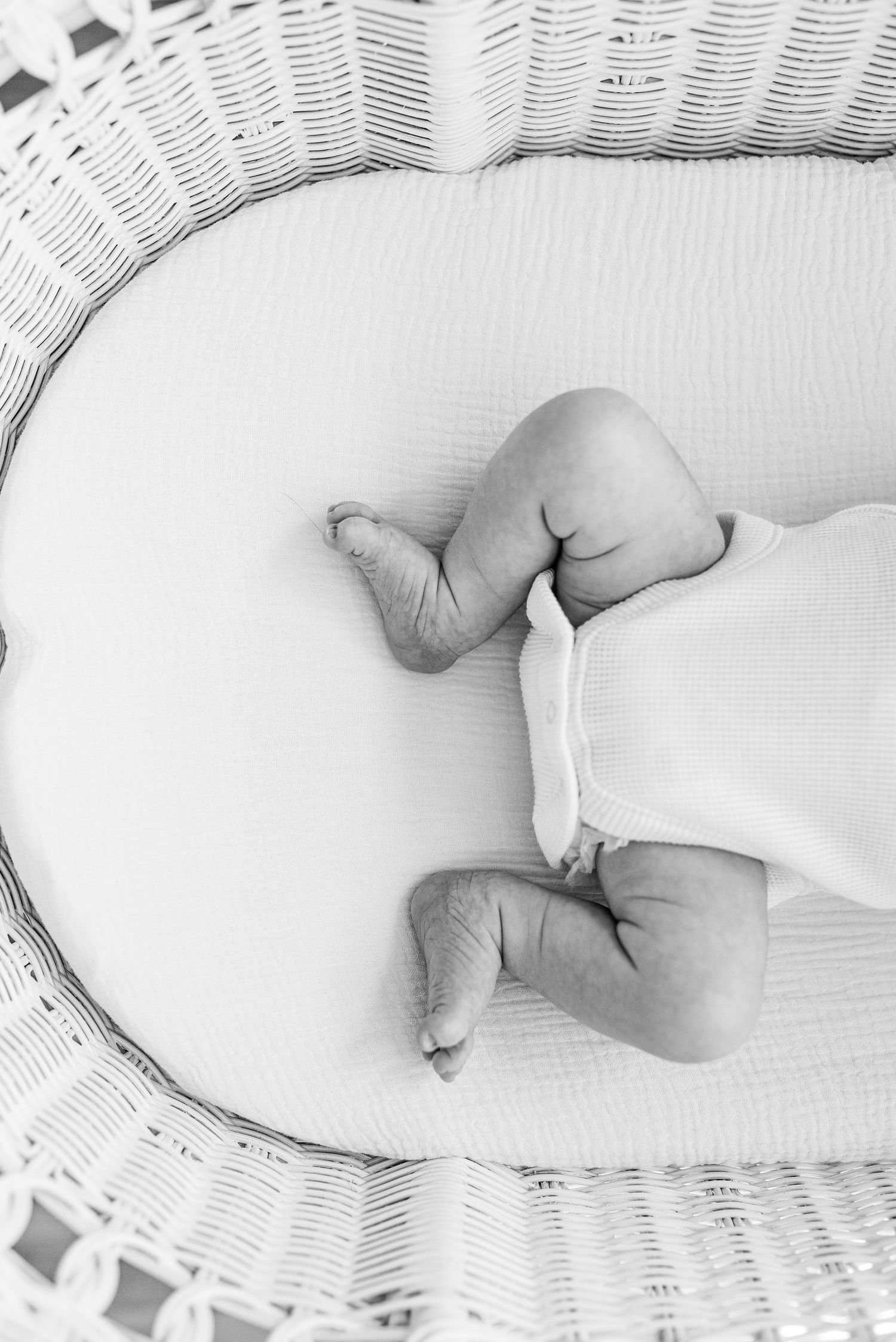 Black and white photo of tiny baby feet and legs resting on white bedding in a wicker bassinet.