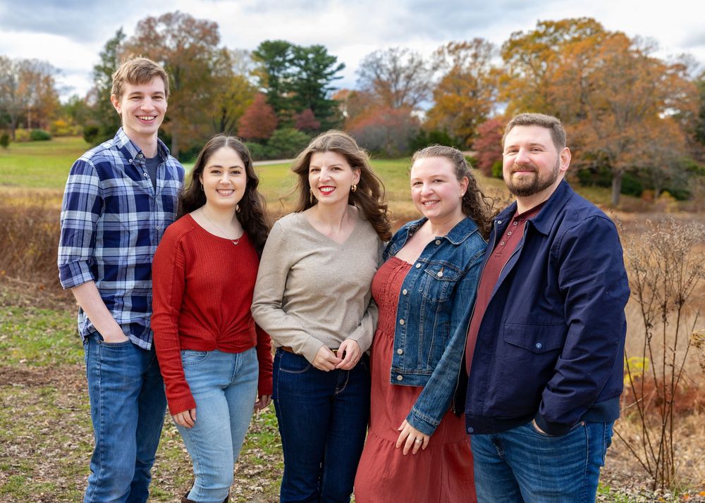 Adult siblings pose together outdoors during autumn with fall foliage and trees visible in the background at Old North Bridge in Concord.
