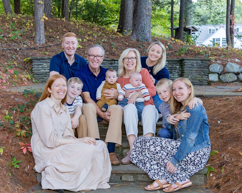 Parents with their adult children and grandchildren pose on steps during an extended family photo shoot surrounded by greenery and trees.
