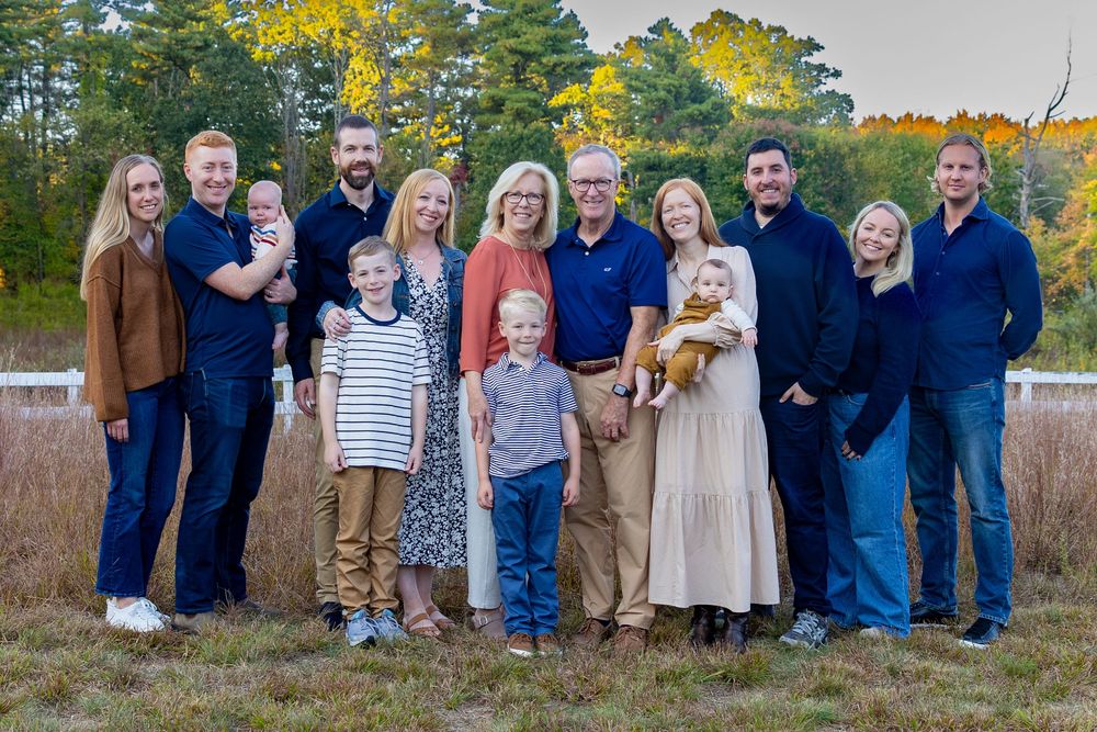 A group of people in coordinated blue and neutral outfits pose together outdoors during autumn pose during an extended family photo shoot.