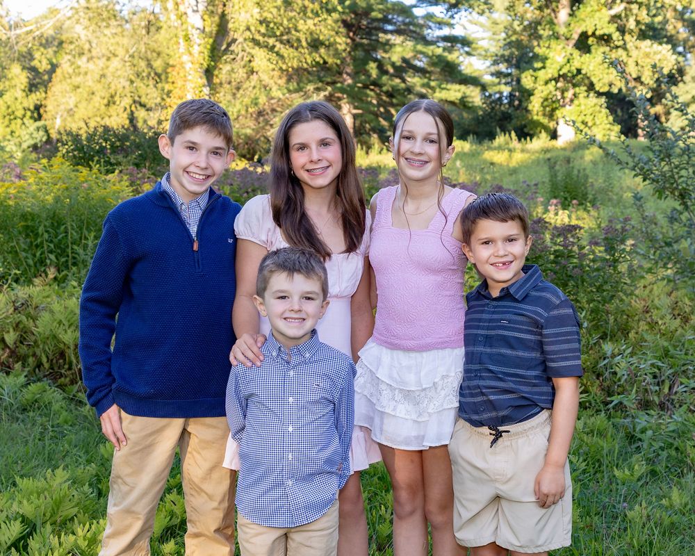 A group of cousins pose together outdoors in casual clothing against a green summer landscape with trees.