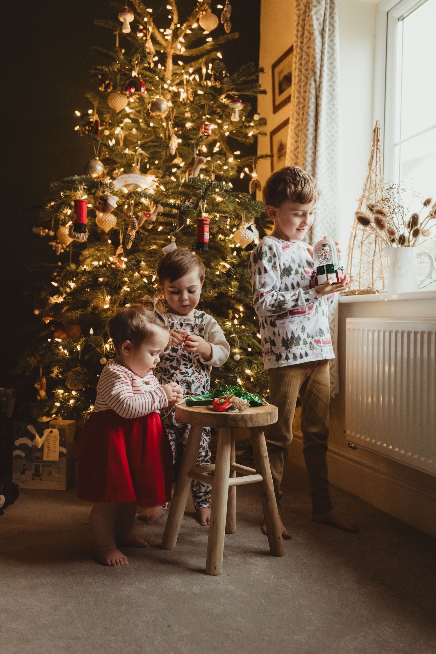 Children gather around a wooden stool near a decorated Christmas tree with twinkling lights in a cozy living room.