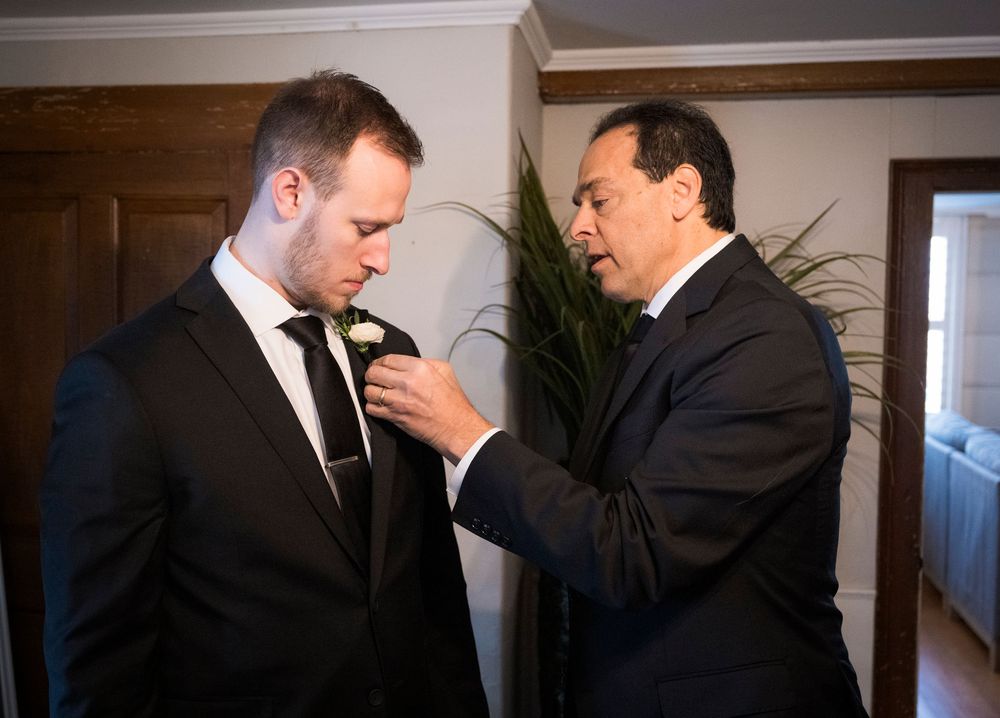 Two men in formal attire adjusting tie and boutonniere during wedding preparations.