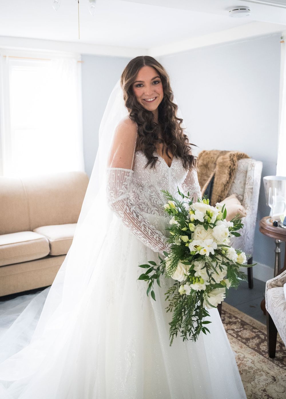 A bride in a white lace wedding dress holds an elegant bouquet of white flowers and greenery in an indoor setting.