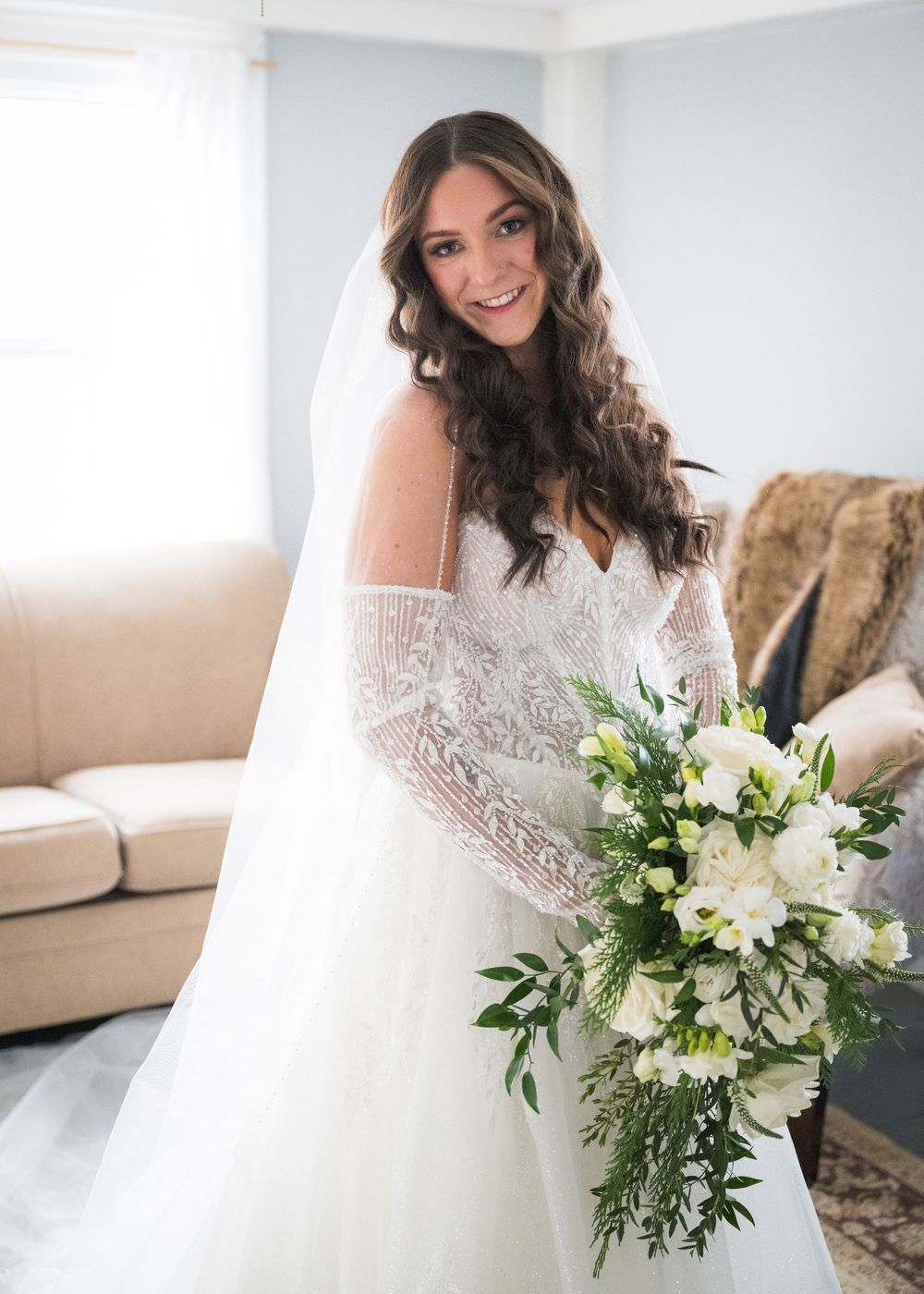 A bride in a lace off-shoulder wedding dress holds a white and green bouquet while standing in a bright room.