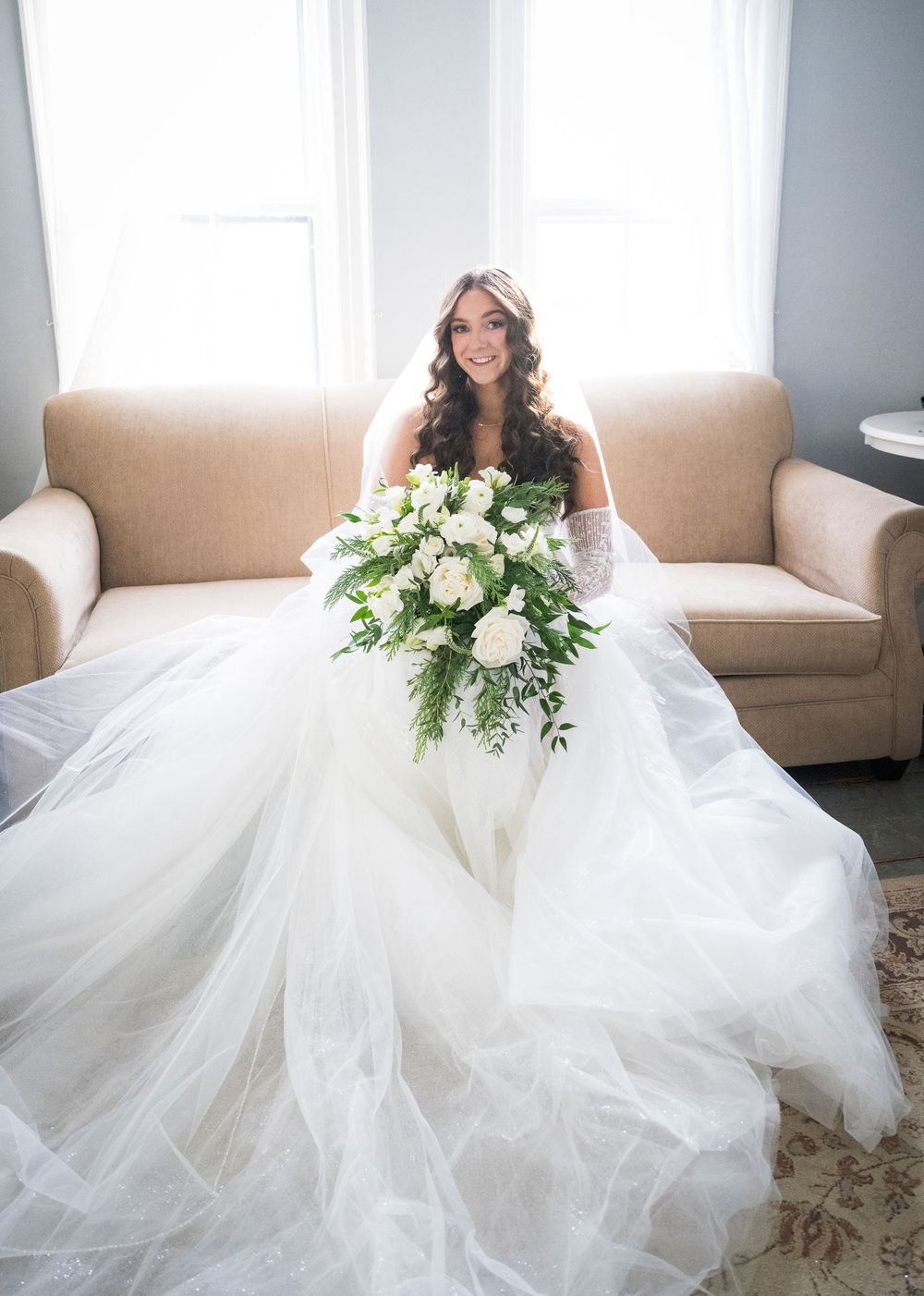 Elegant bridal portrait with flowing white wedding gown and white rose bouquet on beige sofa.