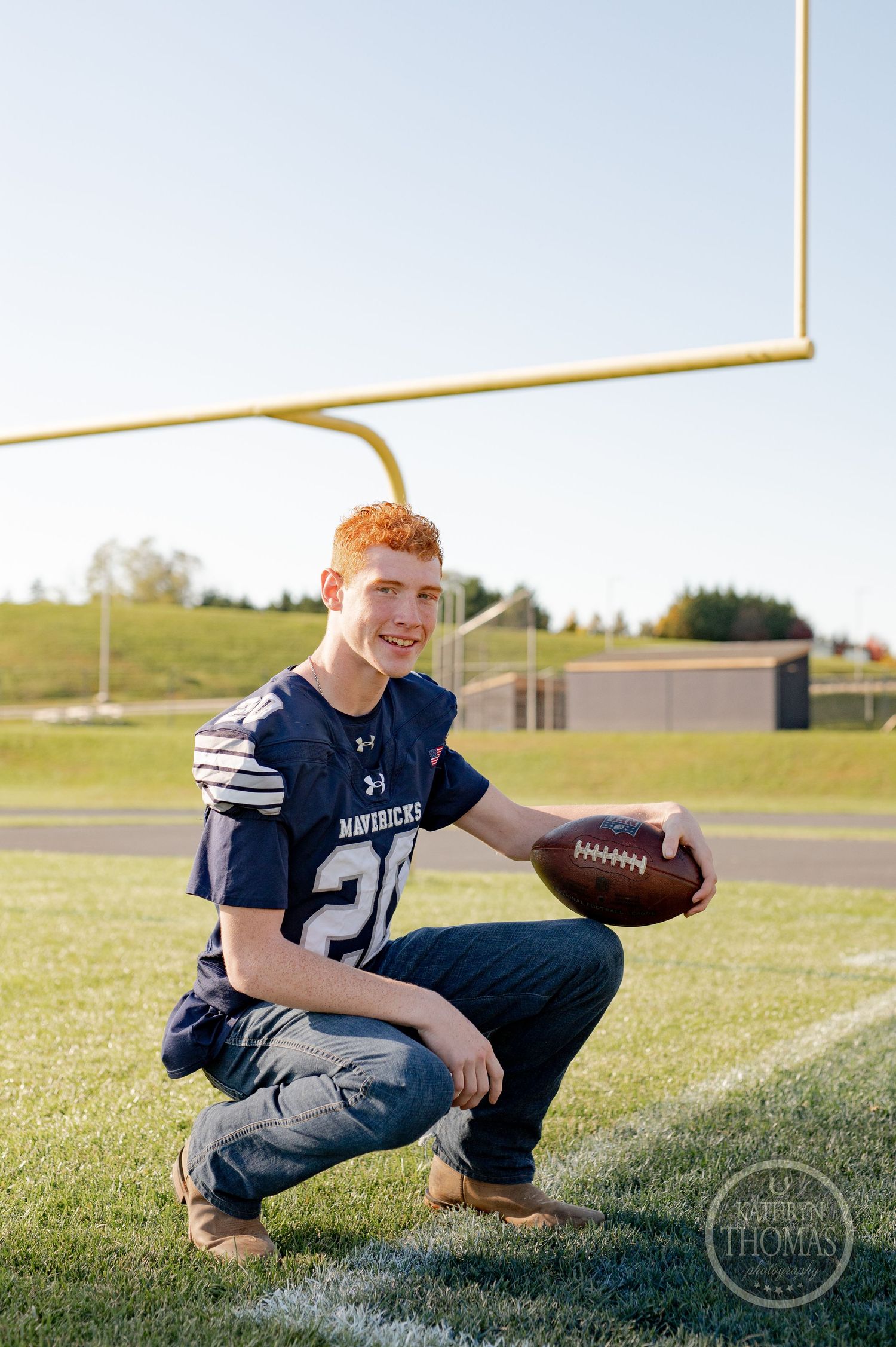 A football player in a navy Mavericks jersey crouches on the field holding a football.
