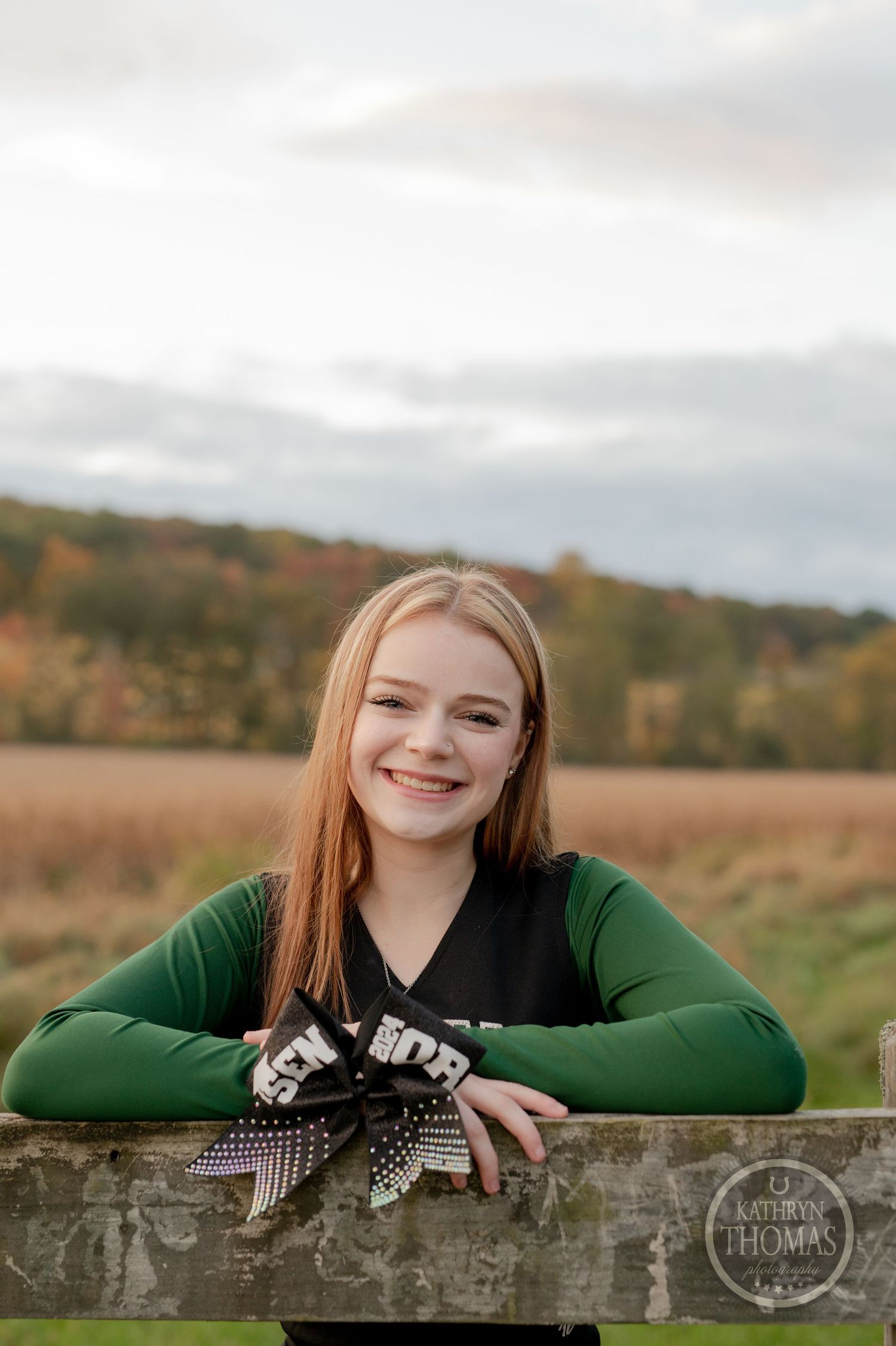 Cheerleader in green and black uniform poses against rustic fence.