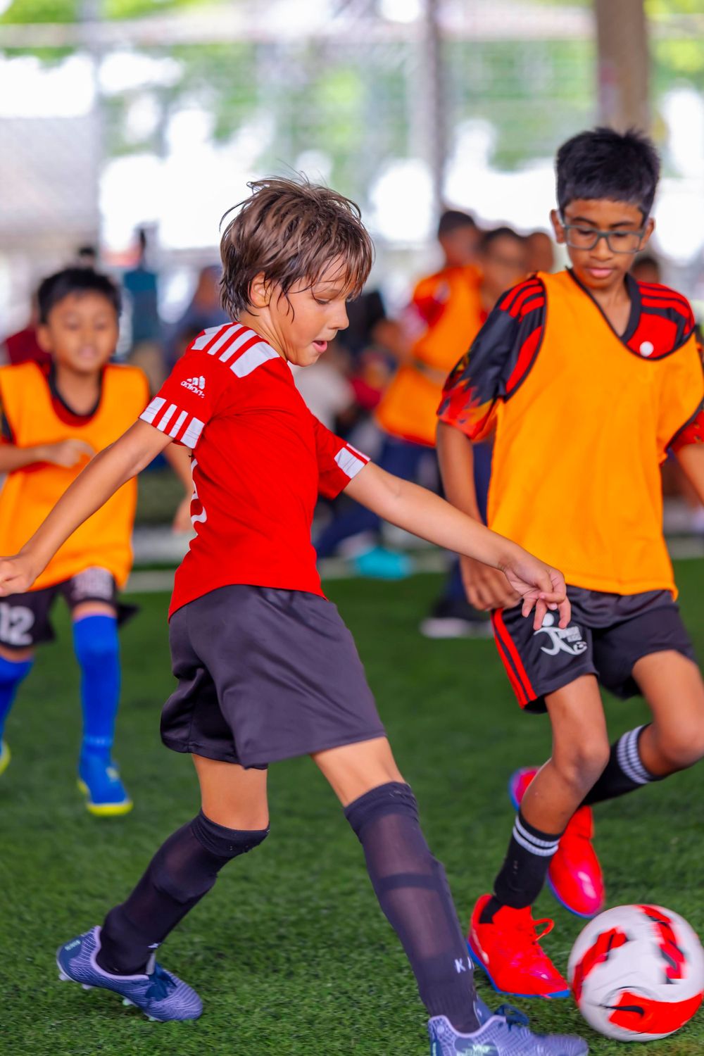 Youth soccer players in red and orange jerseys competing for the ball on artificial turf.