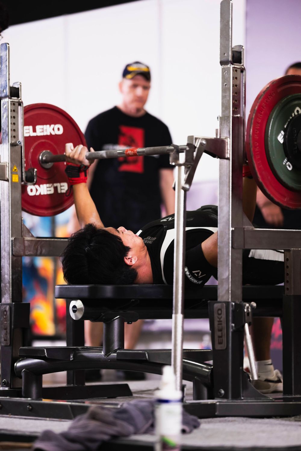 Powerlifter performing a bench press with heavy weights on a barbell while being spotted in a gym setting.