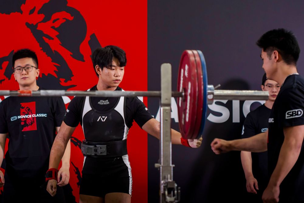 Athletes preparing for weightlifting with loaded barbell in front of red backdrop.