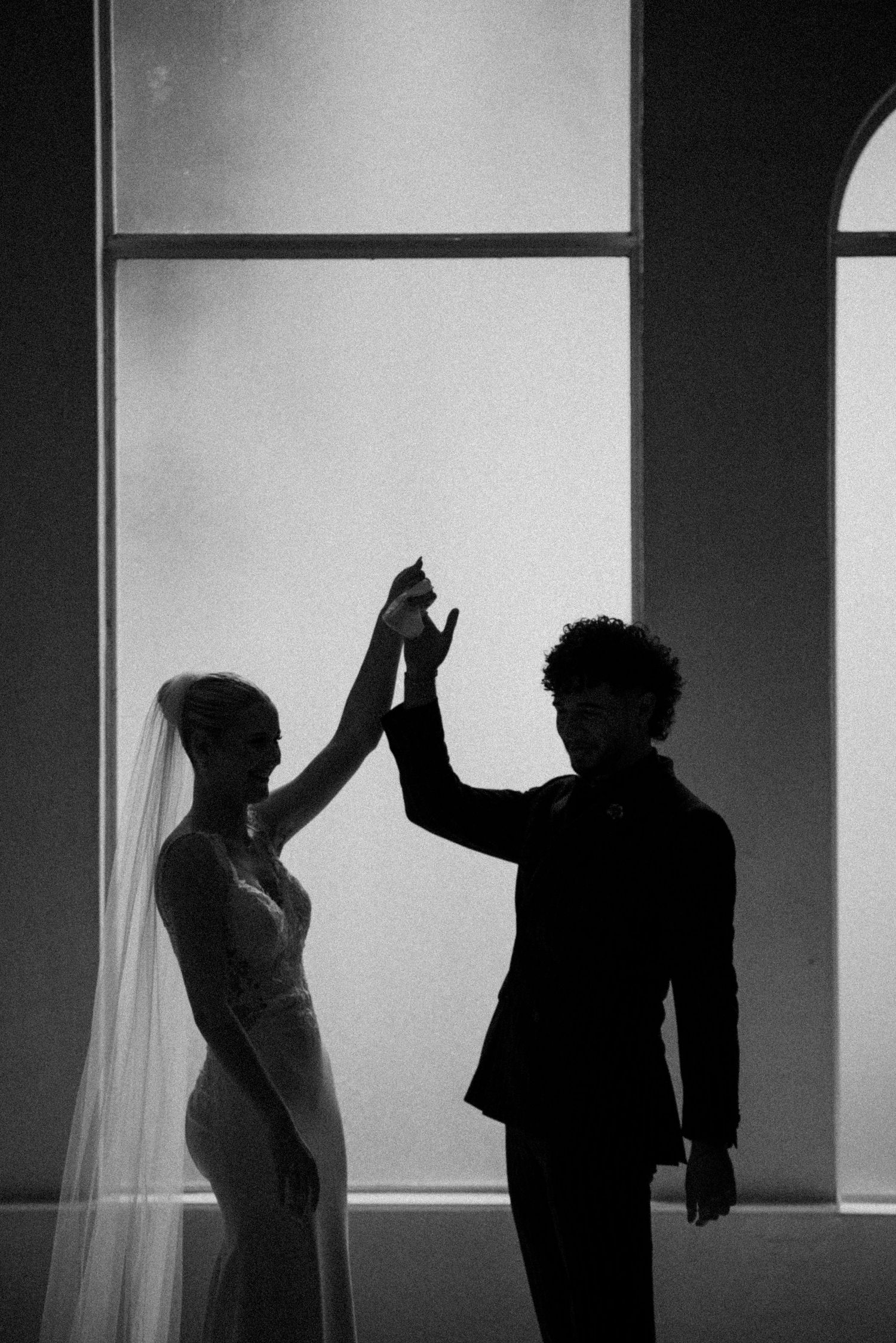 A silhouetted couple raises their hands together in a dramatic black and white wedding photo.