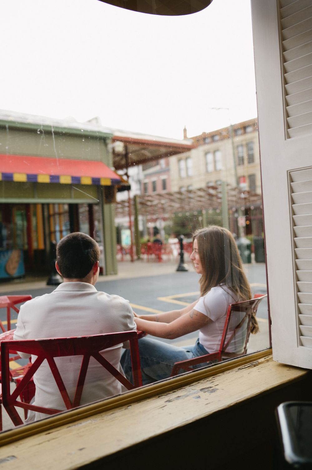 View through window of couple sitting at outdoor cafe in red chairs on city street.