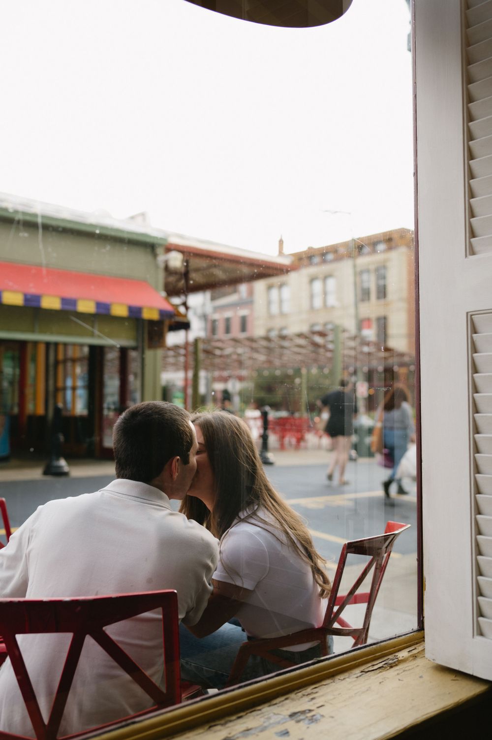 A couple shares an intimate moment at a cafe table by the window overlooking a city street.