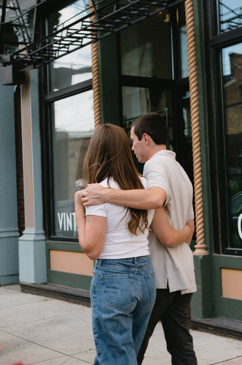 A couple embraces while walking past storefronts on an urban sidewalk.