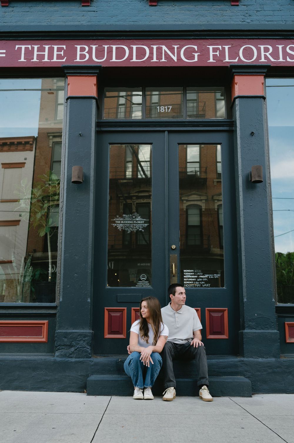 A couple sits on steps outside The Budding Florist shop storefront.