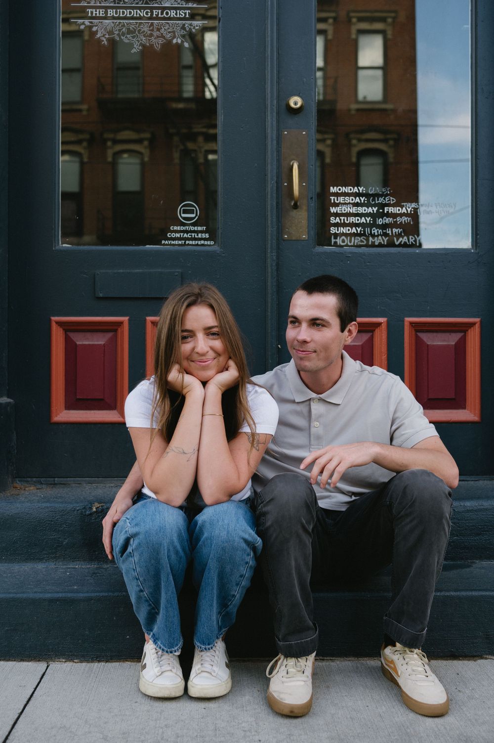 A couple relaxes on steps outside a blue storefront with red accents.