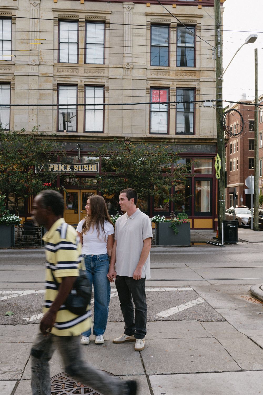 Couple standing on urban street corner with historic brick building and pedestrian walking past.