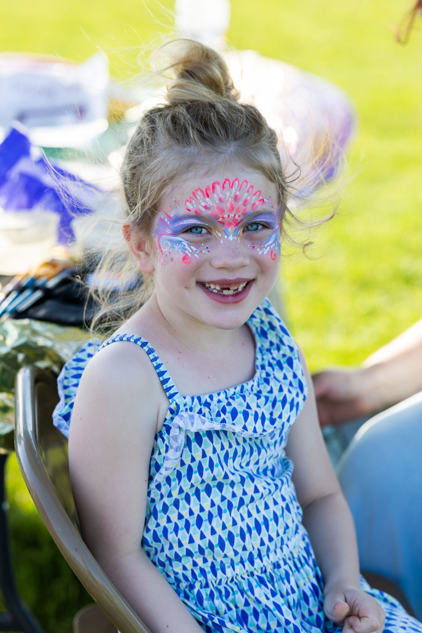 Child in blue dress shows off colorful face paint design at outdoor event.