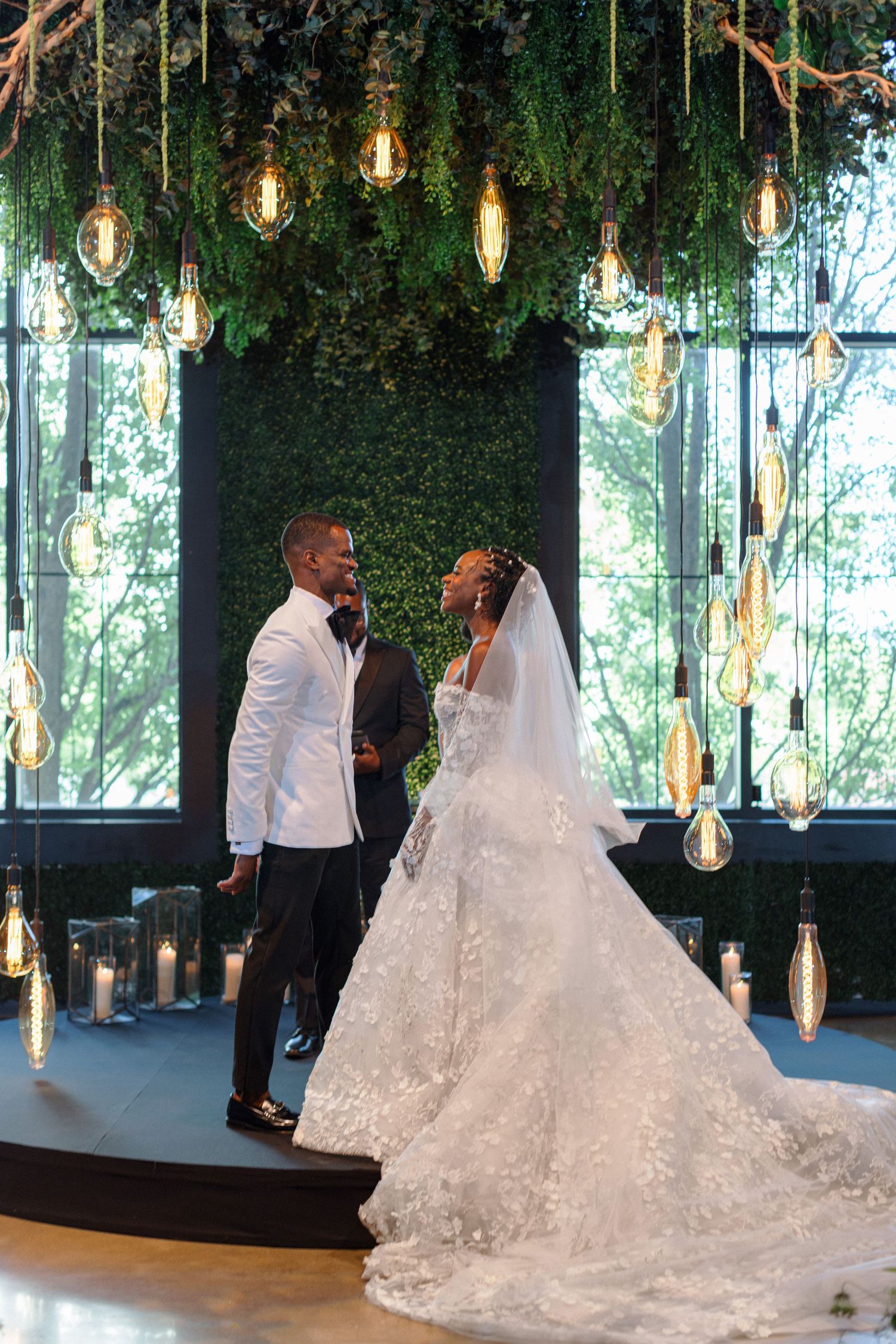 A wedding ceremony under a canopy of hanging Edison bulbs and greenery in a modern venue.