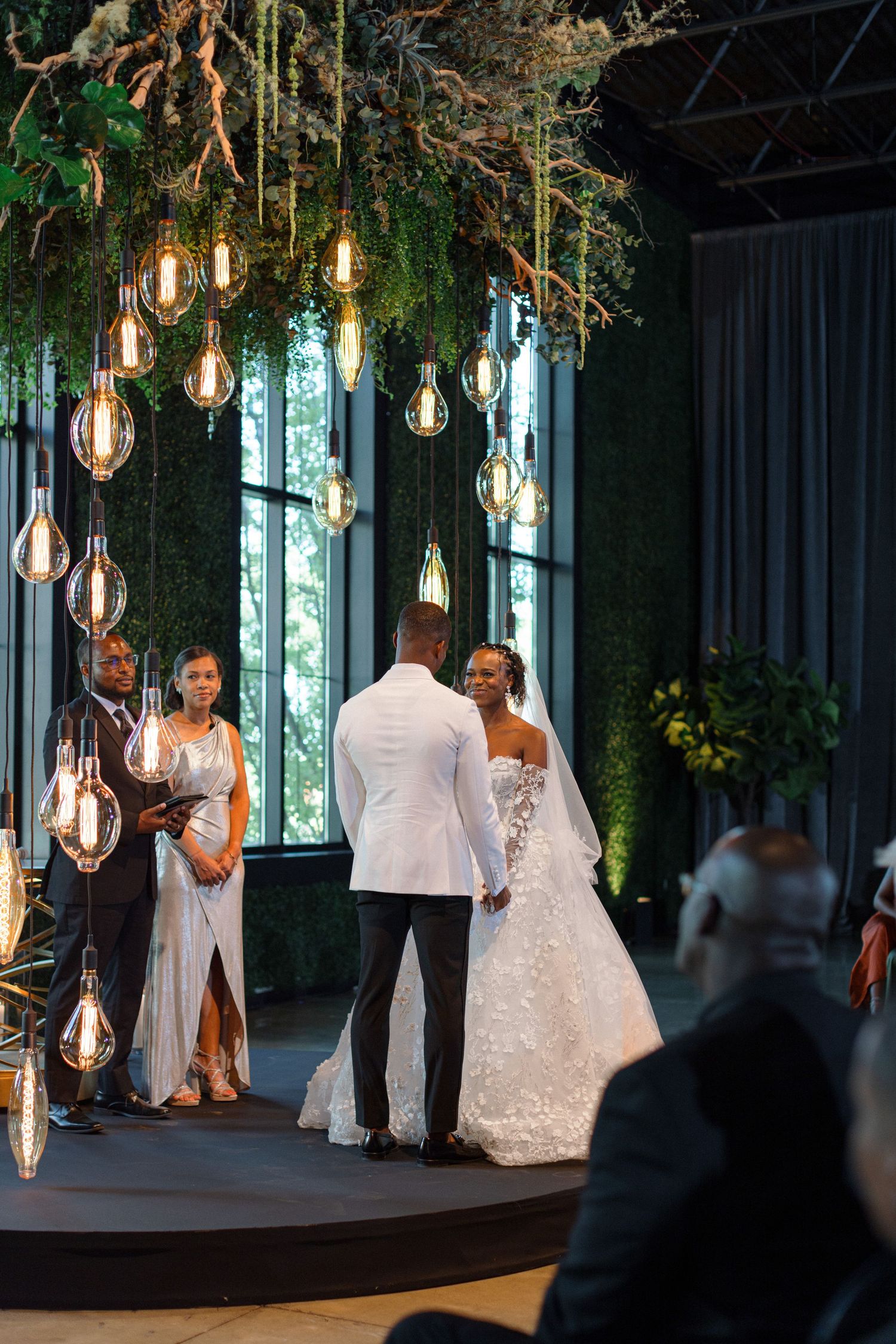A wedding ceremony in progress under suspended Edison bulbs with lush greenery and floor-to-ceiling windows.