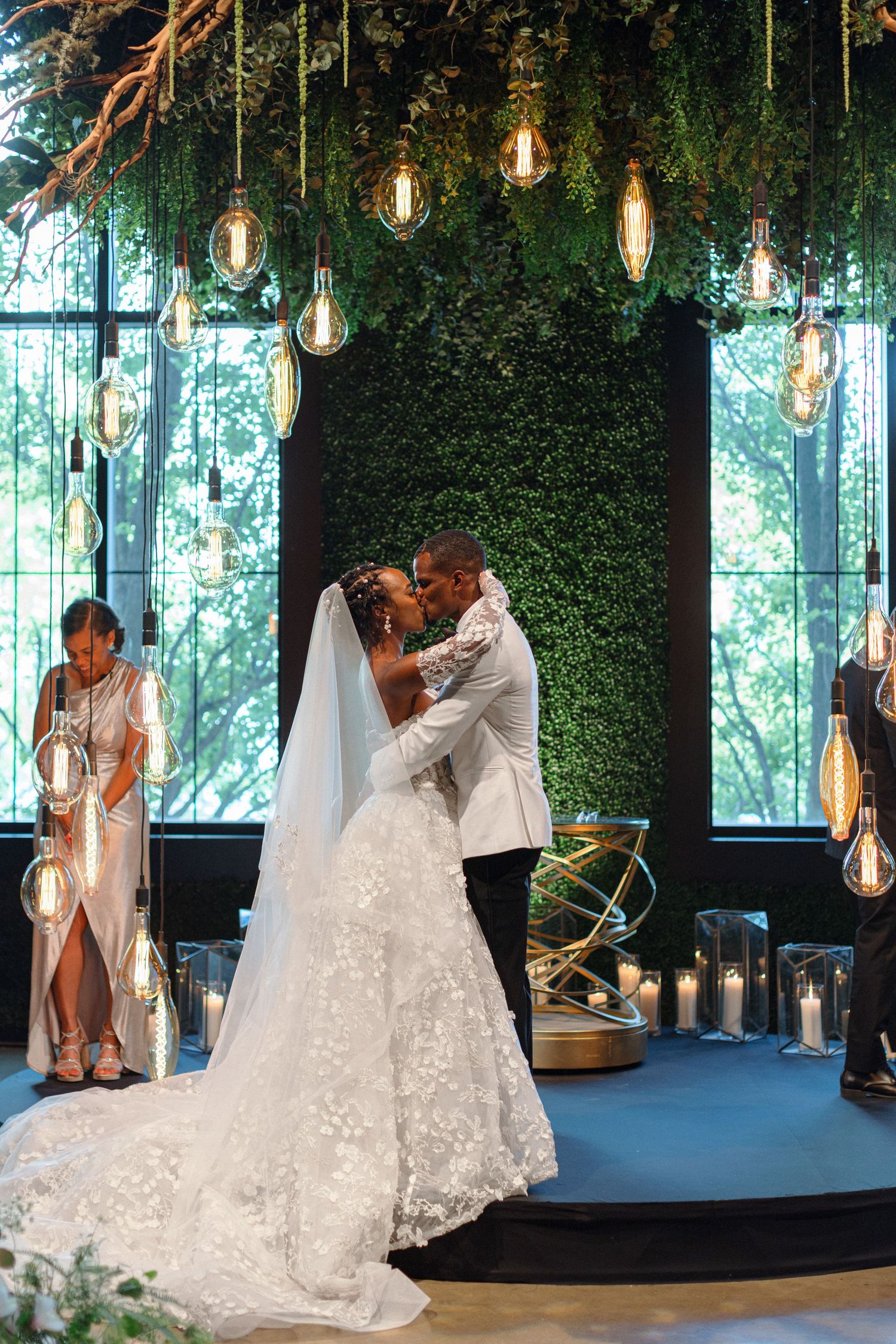 A romantic wedding moment under suspended Edison bulbs and a wall of greenery.