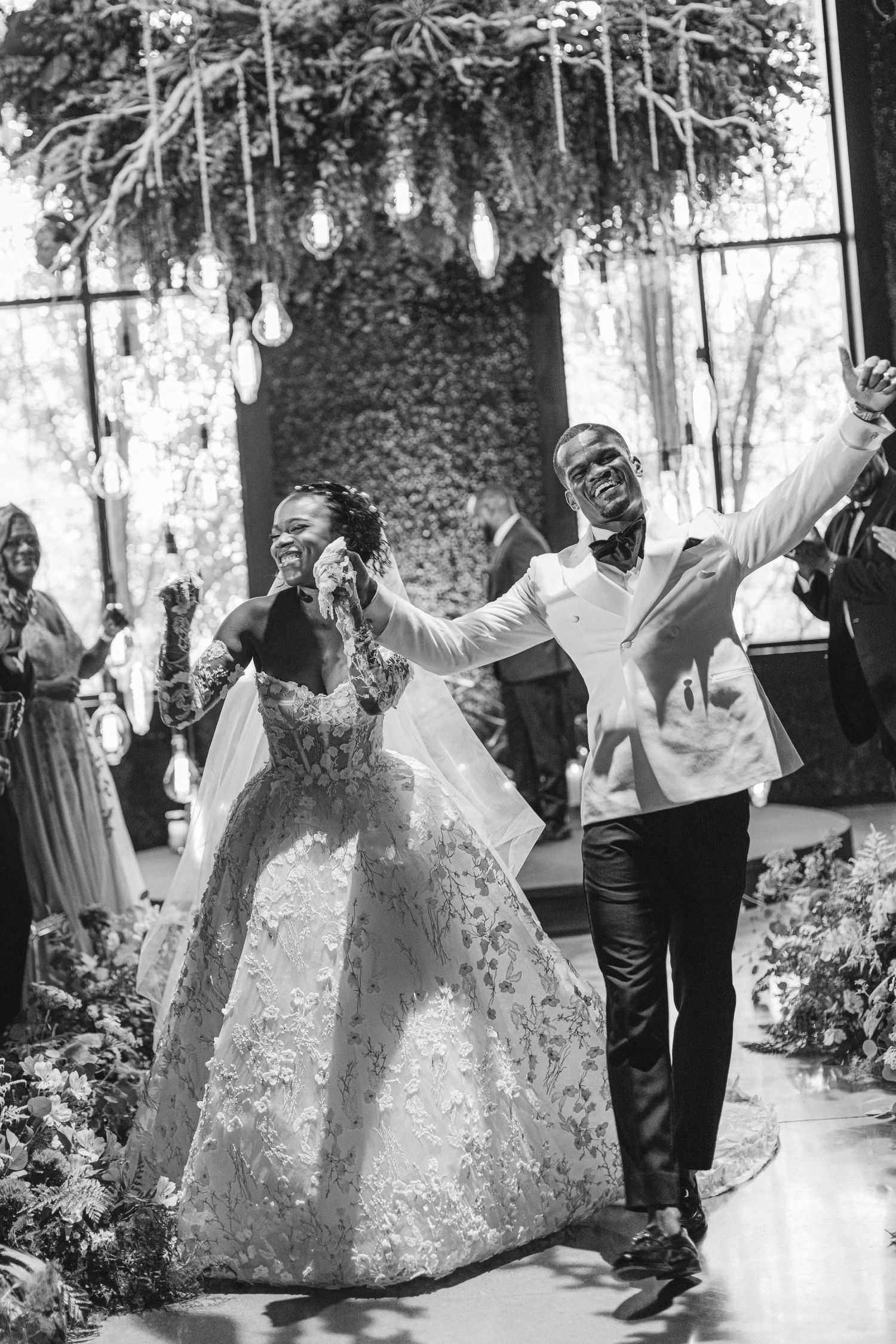 A joyful wedding celebration captured in black and white under hanging lights and greenery.