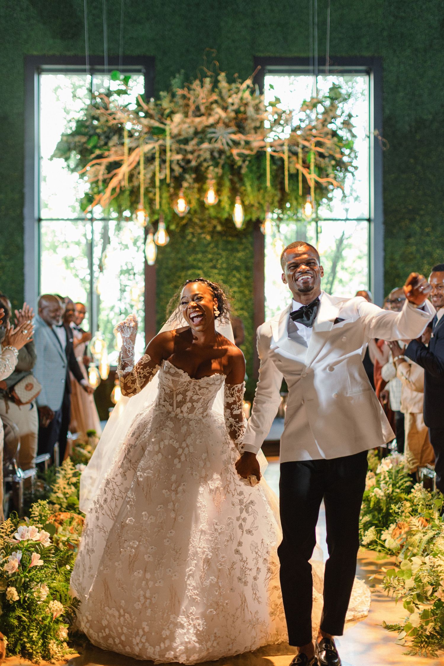 A celebratory wedding recessional under hanging Edison bulbs and floral arrangements.