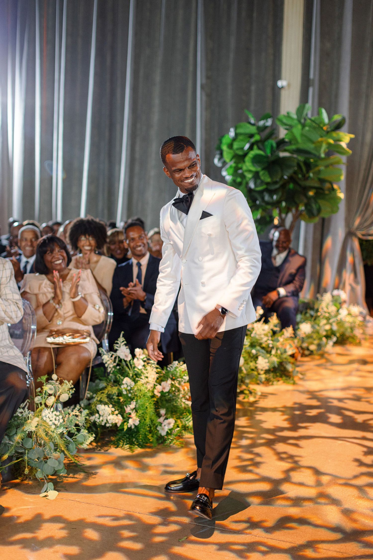 Groom in white tuxedo jacket walks down the aisle with animated expression during wedding ceremony.